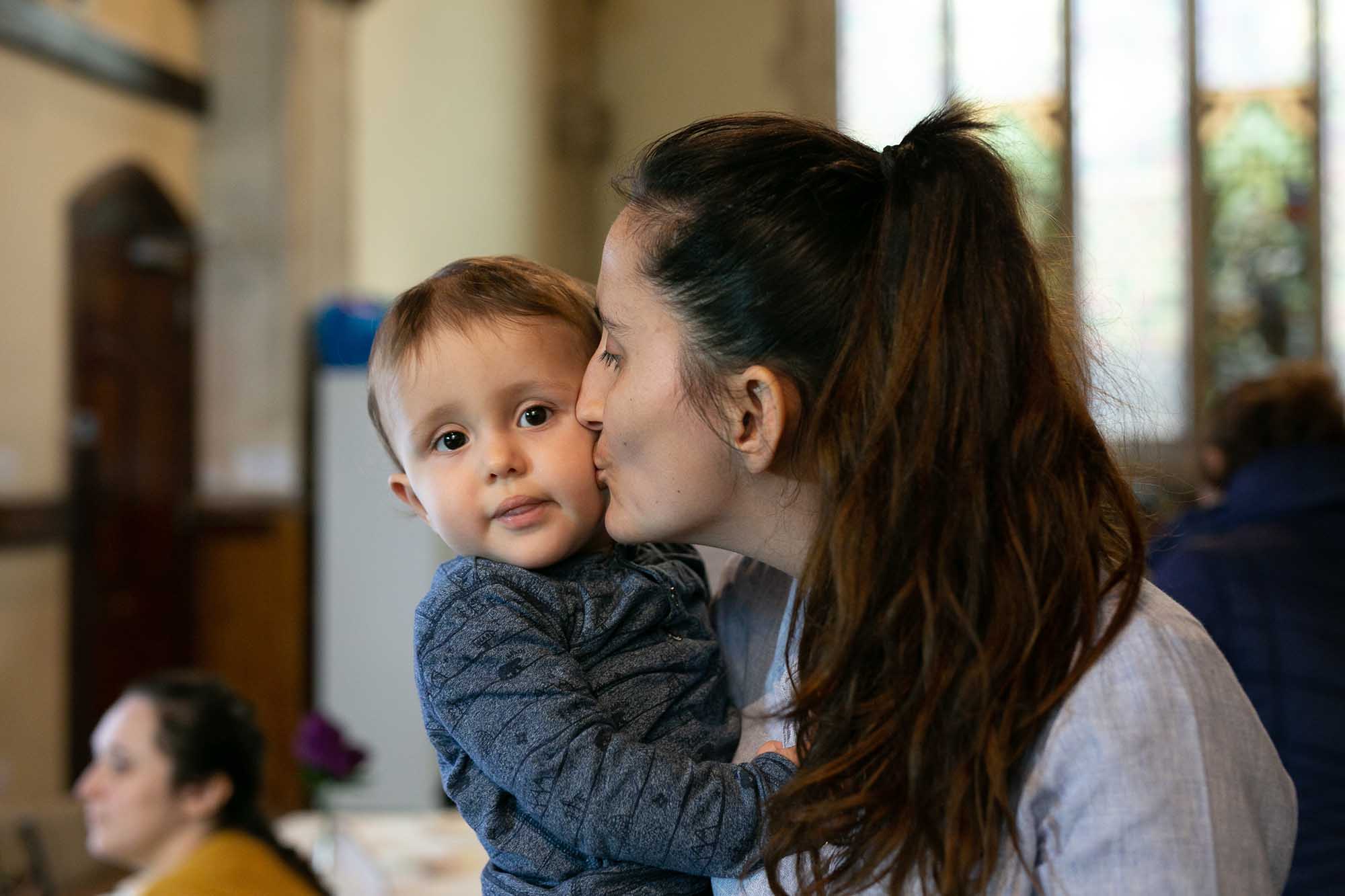 A mother with dark brown hair kisses her infant on the cheek.