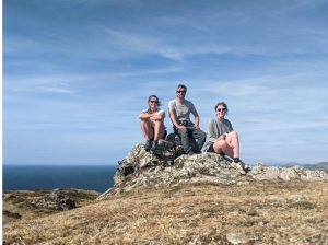 A photograph of a group of three people sit on a mountain trig point.