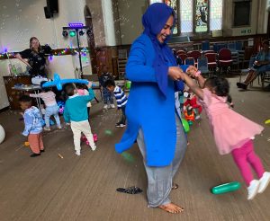 A woman in a blue scarf and tunic spins a child around during a play session