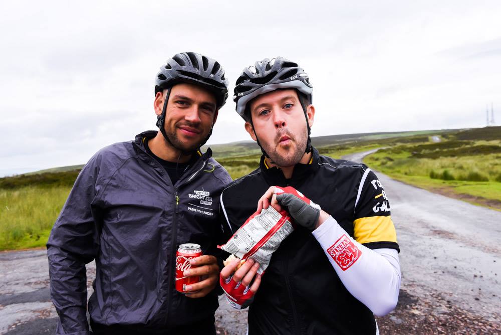 Two cyclists in helmets raise drinks on a moorland road
