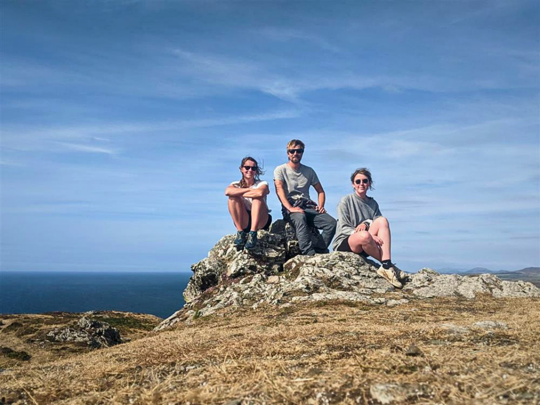 Three people sit on a rock at the top of a mountain smiling at the camera
