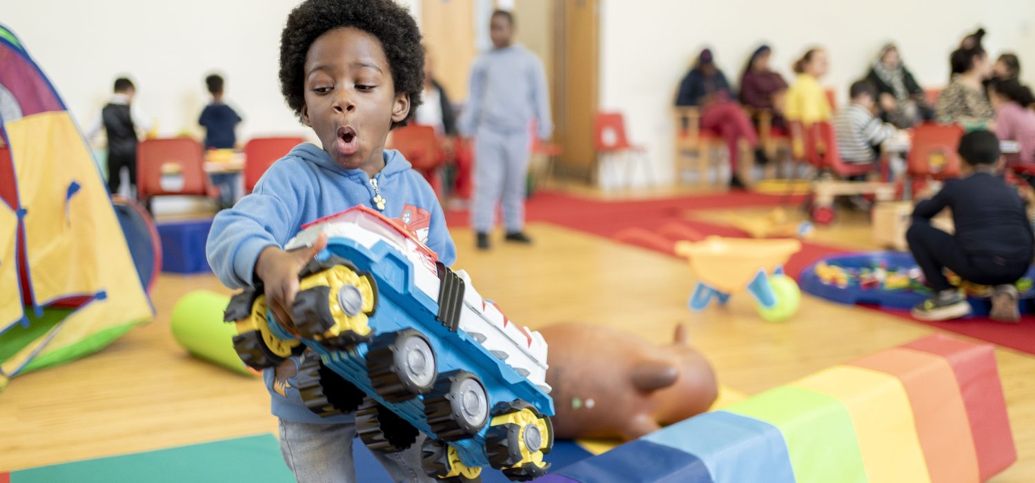 A young boy plays with a large toy truck in a brightly lit room with rainbow mats. Other children and toys are blurred in background.
