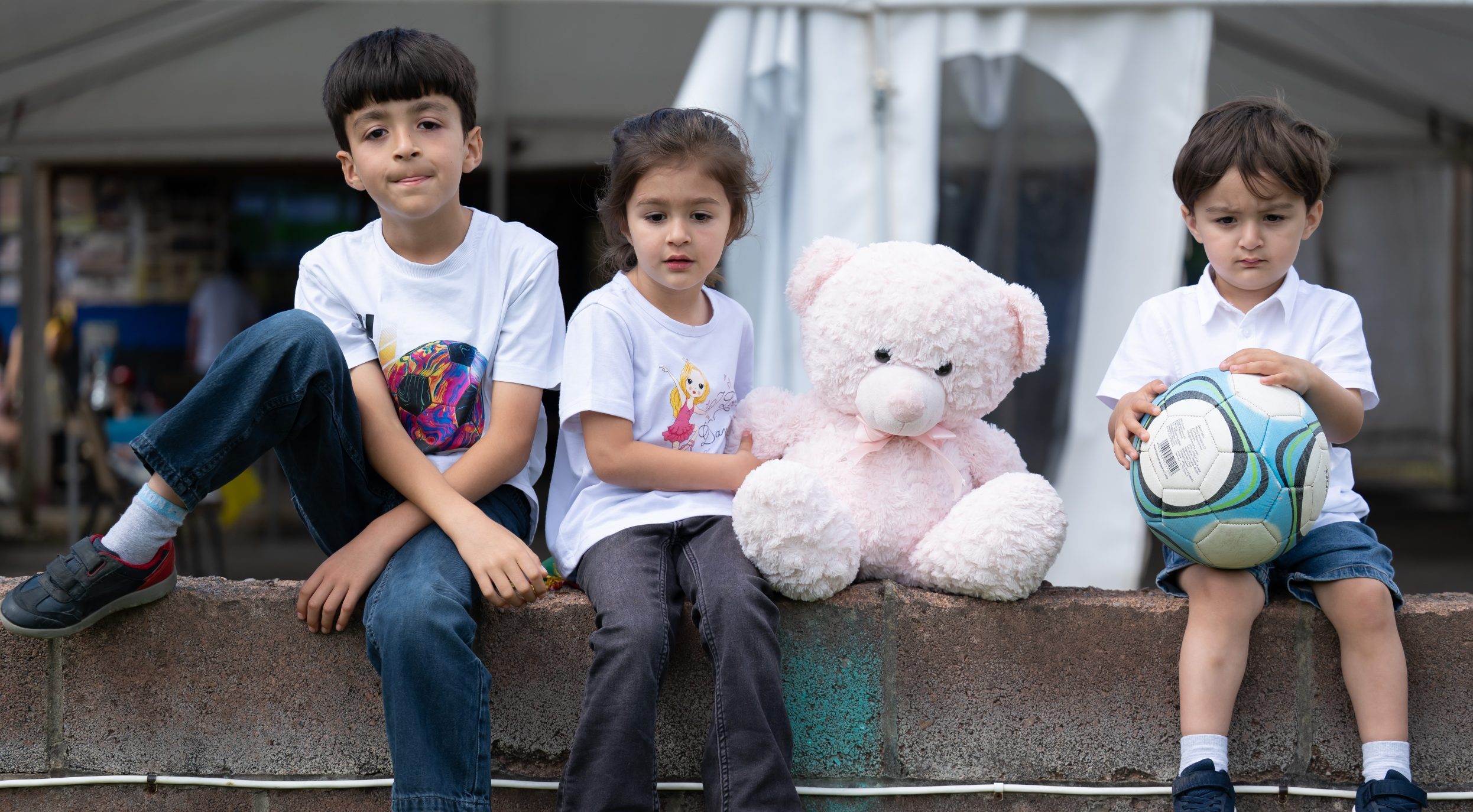 Three children sit next to a large pink stuffed bear on a ledge. One holds a football.