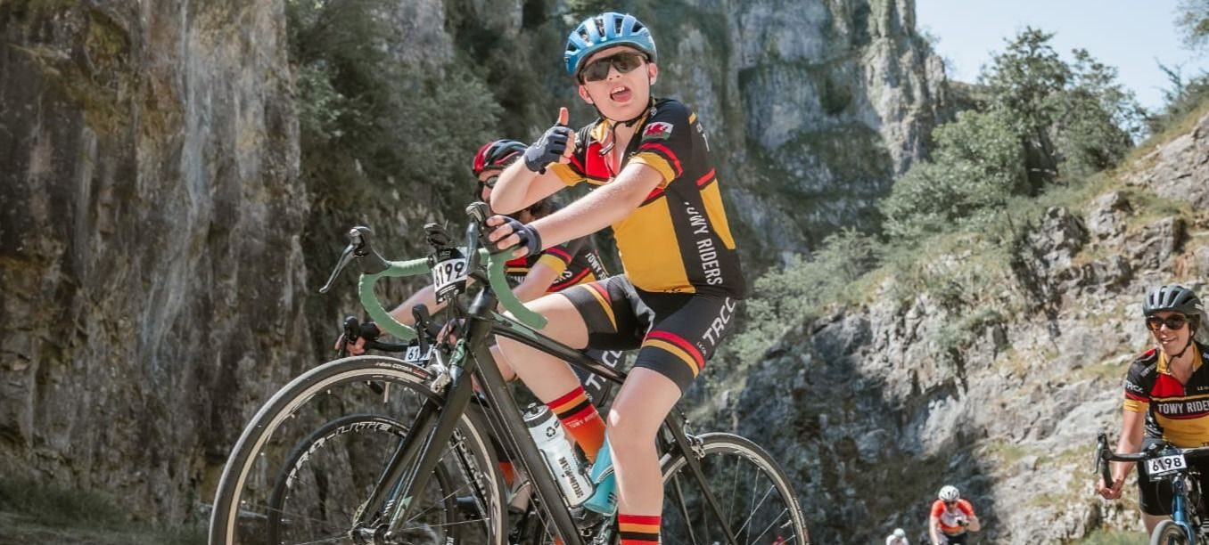 A young cyclist gives a thumbs up while riding up a mountain road