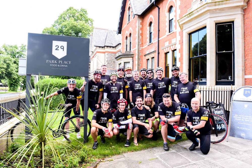 The Tour de Galles riders in matching black jerseys pose with their bikes outside a red-brick building